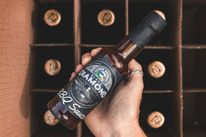 A person holds a bottle of Black Diamond BBQ Sauce, with a box of beers underneath 