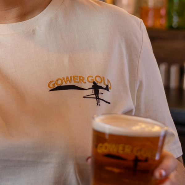 Closeup of a person wearing a Gower Gold T-shirt holding a Gower Gold pint glass