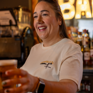 Woman in a bar holding a drink, wearing a white Gower Gold T-shirt with a logo to the left chest 