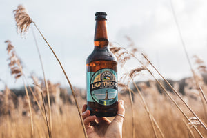 A person holding a bottle of Lighthouse Welsh Lager beer in a field with tall grasses.