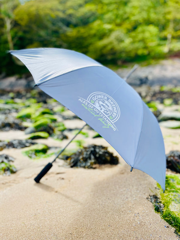 A stylish grey walking umbrella with the Gower Brewery logo printed on it, standing upright on a rocky beach surface for display